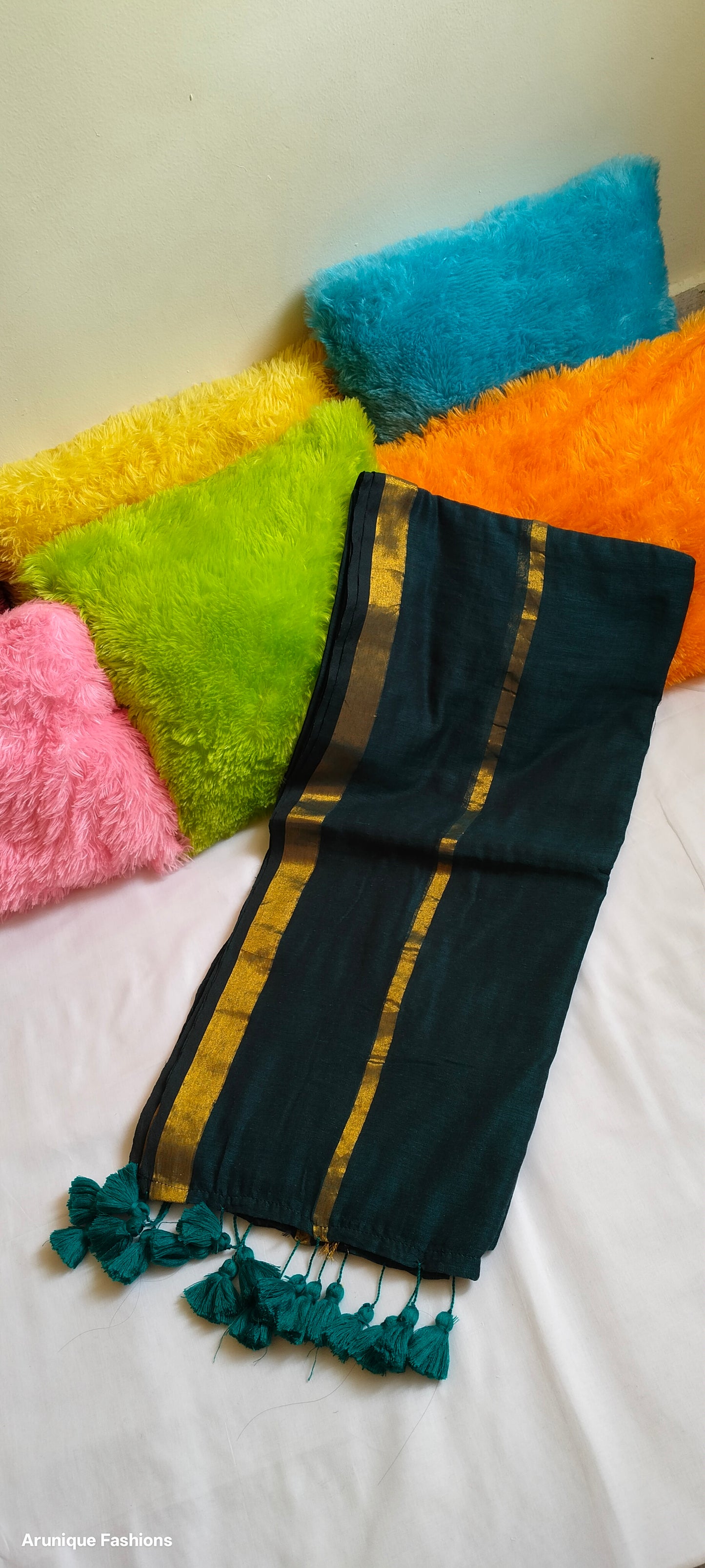 Emerald green double zari mul cotton saree with golden borders and hand-tied green tassels displayed folded with colorful textured fabrics